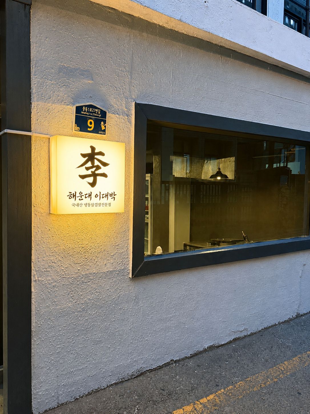 해운대 이대박 매장 외관. 회색 벽면에 따뜻한 노란빛 사각 간판이 켜져 있고, 옆의 긴 창 너머로 매장 안이 보인다.