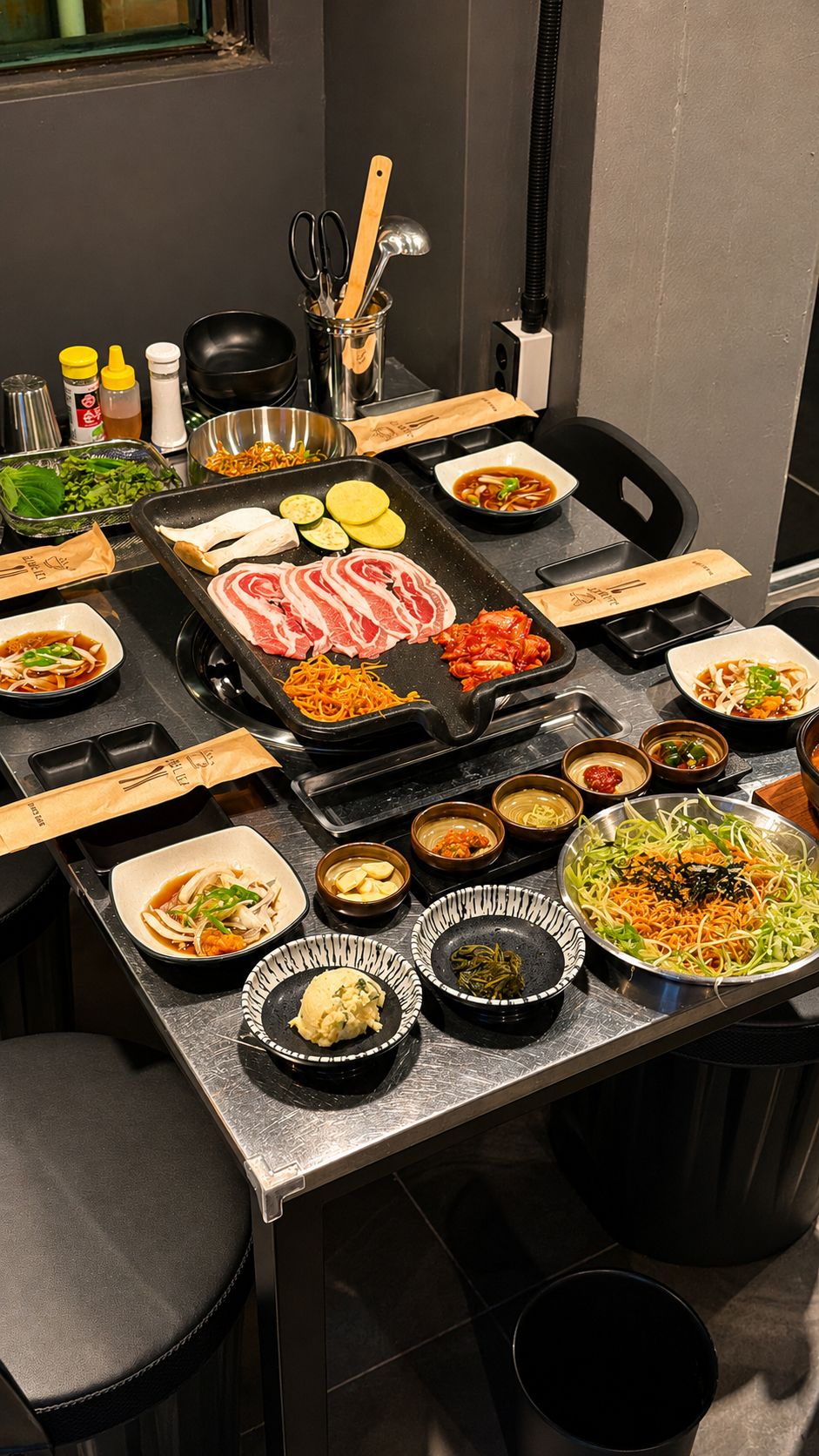 스테인리스 테이블 위 철판에 올린 냉동삼겹살과 김치, 콩나물, 새송이버섯, 애호박과 고구마, 그리고 여러 가지 밑반찬.